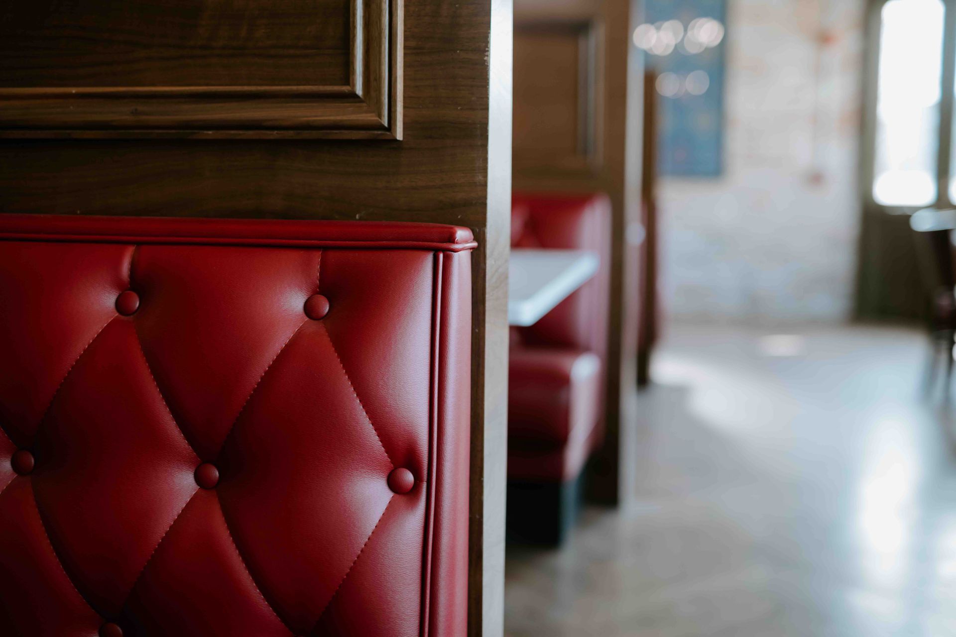 Close-up of a tufted red leather booth seat in a restaurant, with a wooden partition and a blurred dining area behind.