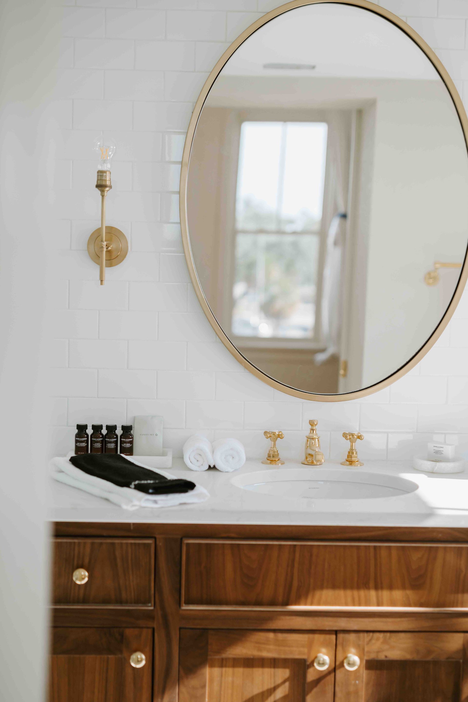 A bathroom vanity with a wood cabinet, white countertop, oval gold-framed mirror, and brass sconce on tiled wall.