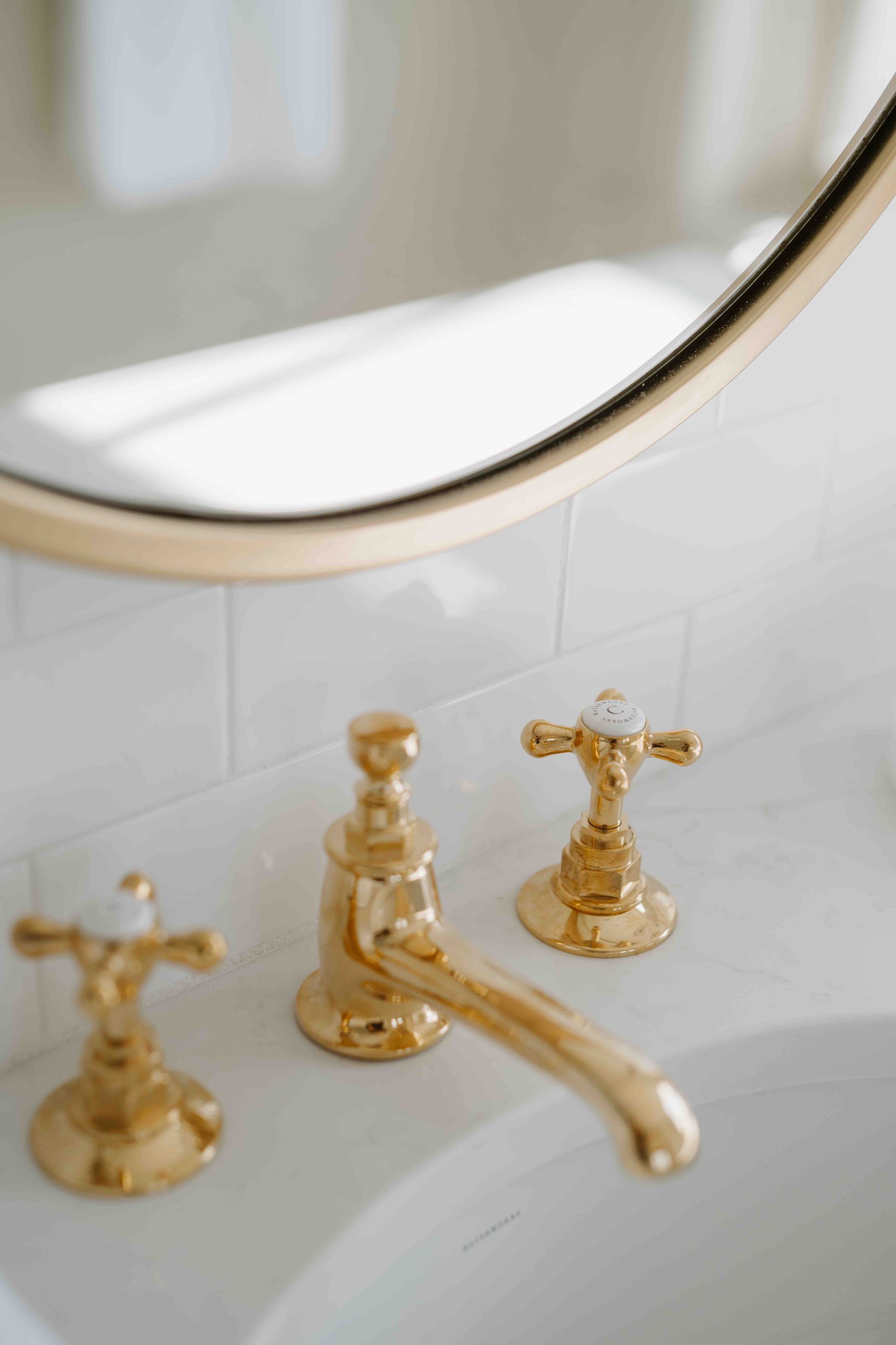 A brass bathroom faucet and handles set against white tiled walls, with a mirror reflected above.