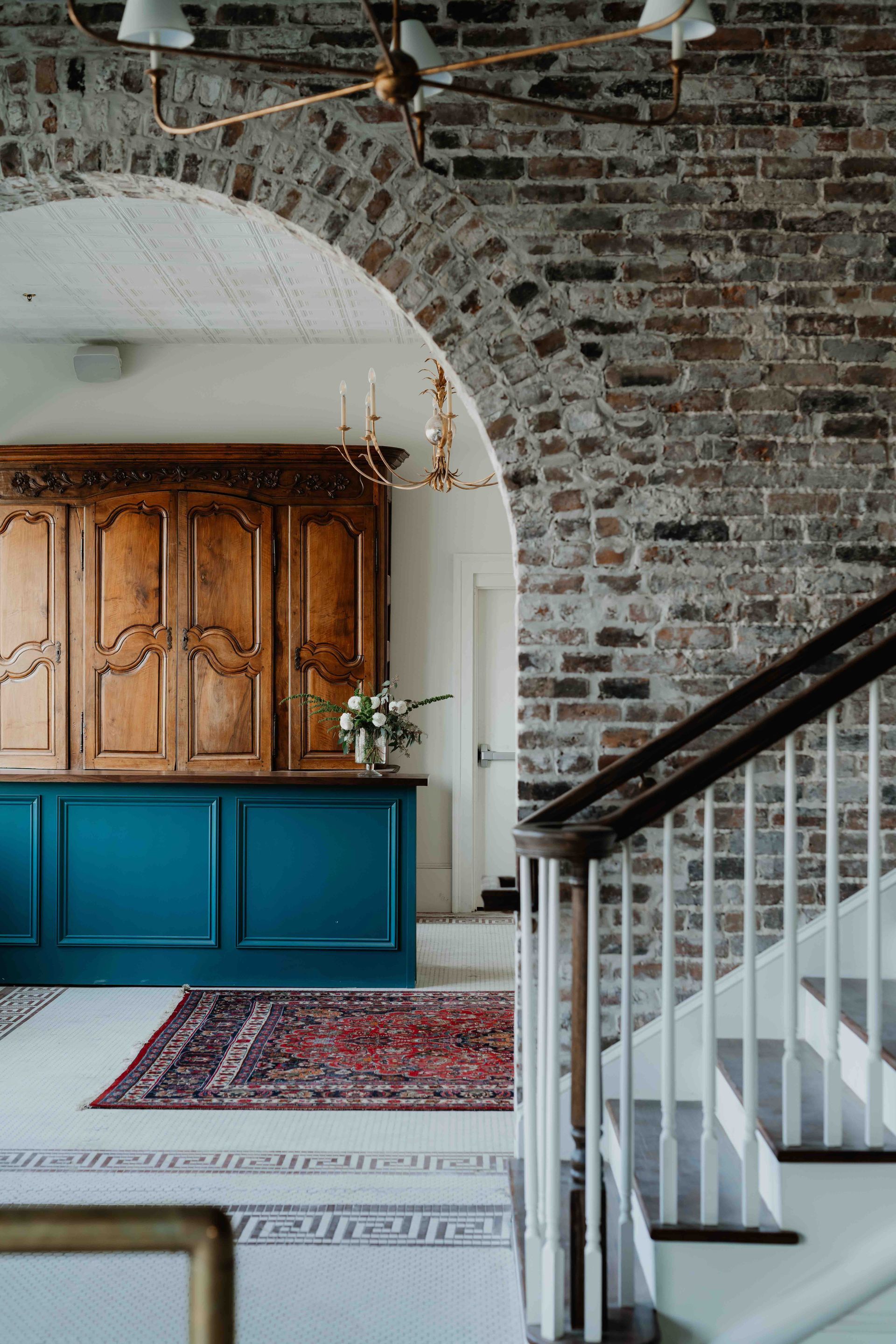 A foyer features a brick archway, a teal cabinet base with a wooden hutch, a patterned rug, and a white staircase.