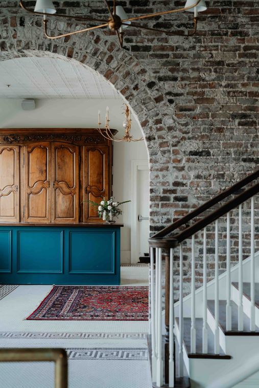 A foyer features a brick archway, a teal cabinet base with a wooden hutch, a patterned rug, and a white staircase.