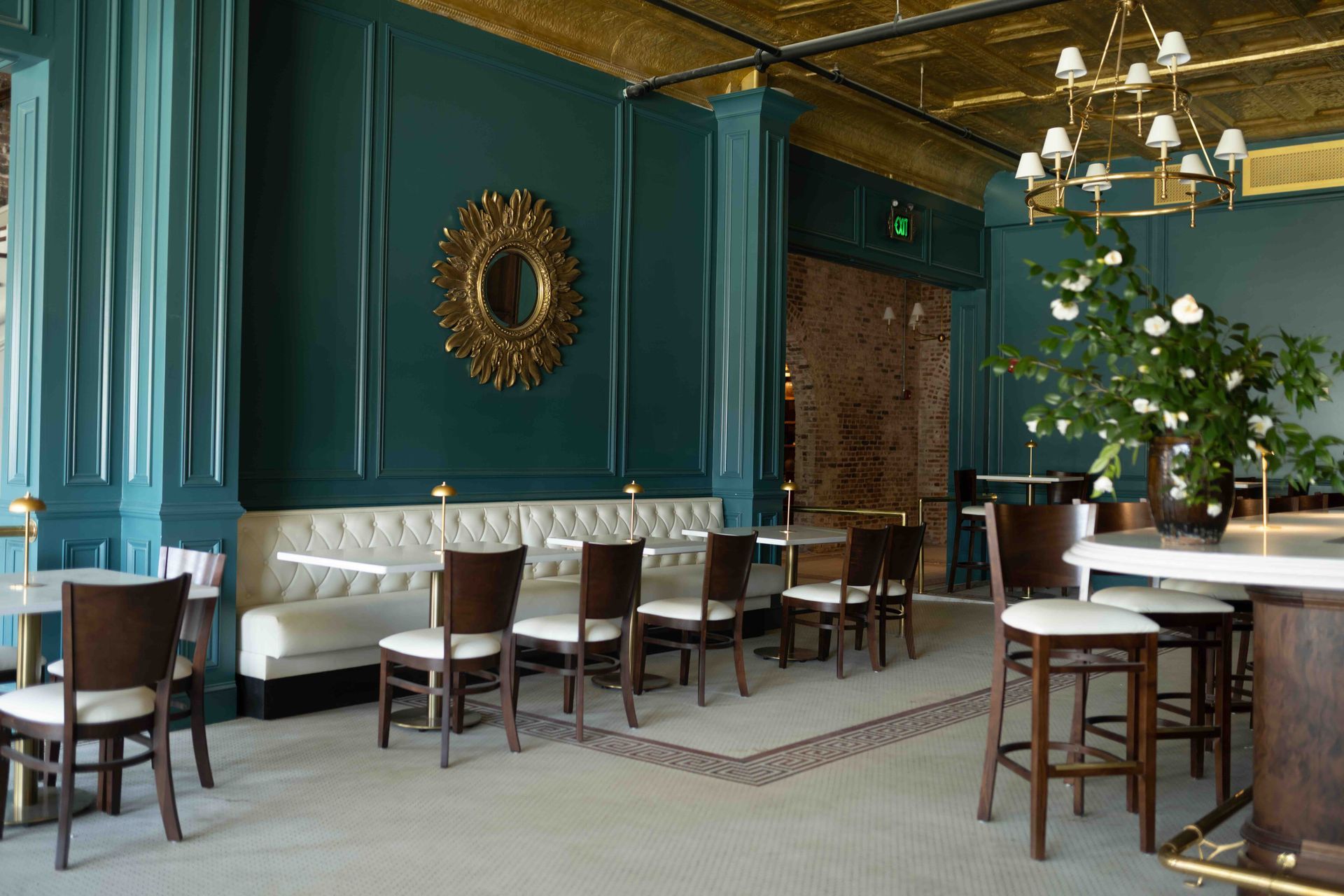 A restaurant interior with teal walls, a white tufted booth, dark wood chairs, a gold ceiling, and a chandelier.
