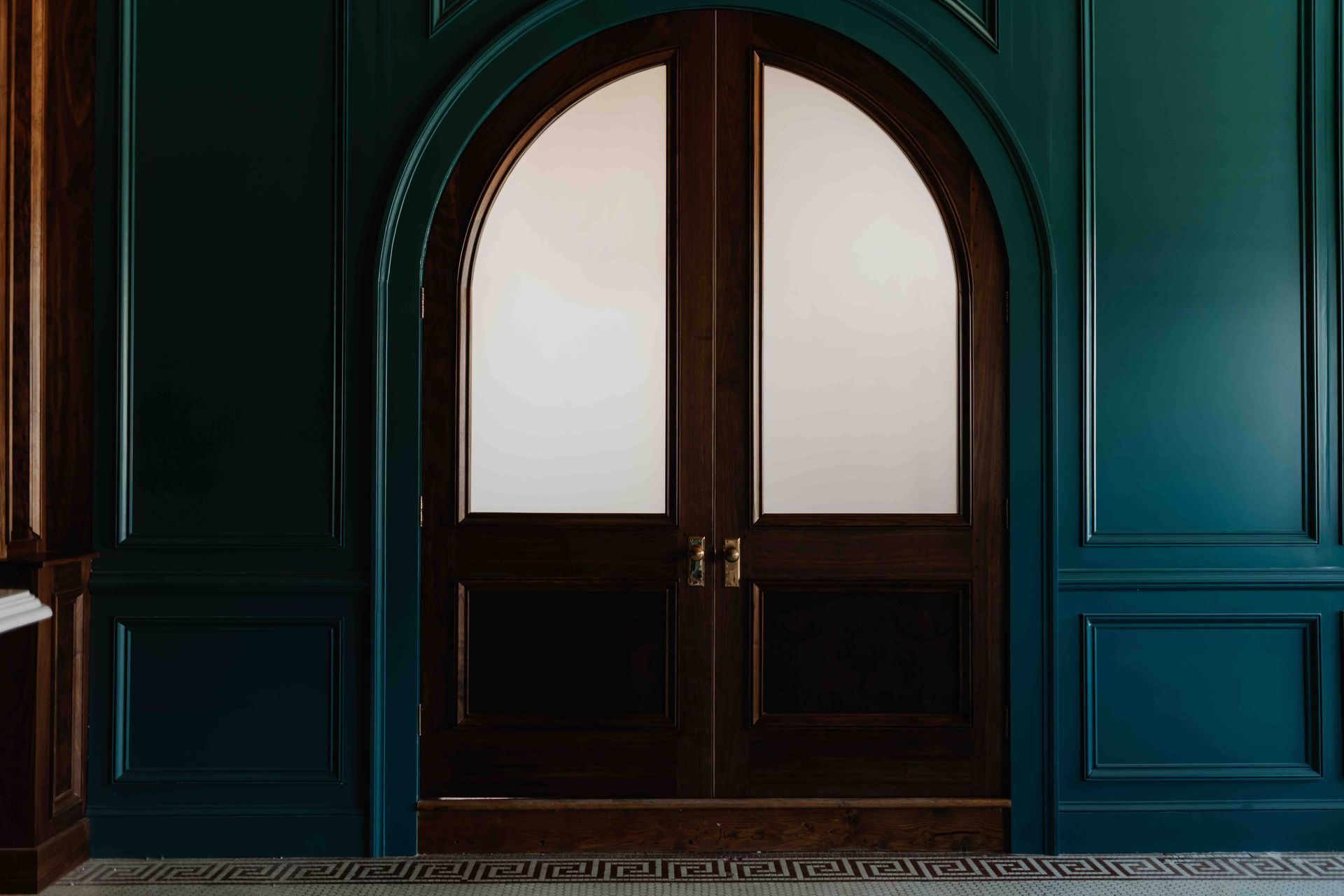 Double dark wood doors with frosted glass panels set in an arched frame against deep green, paneled walls.