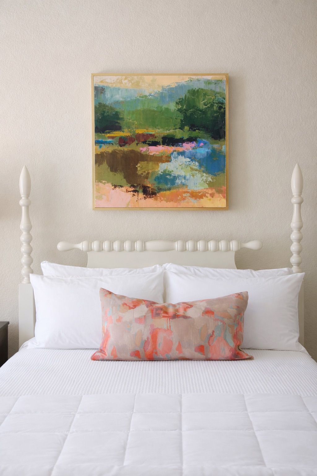 A white spindle bed with a light-colored abstract landscape art piece mounted above it on a textured, light beige wall.