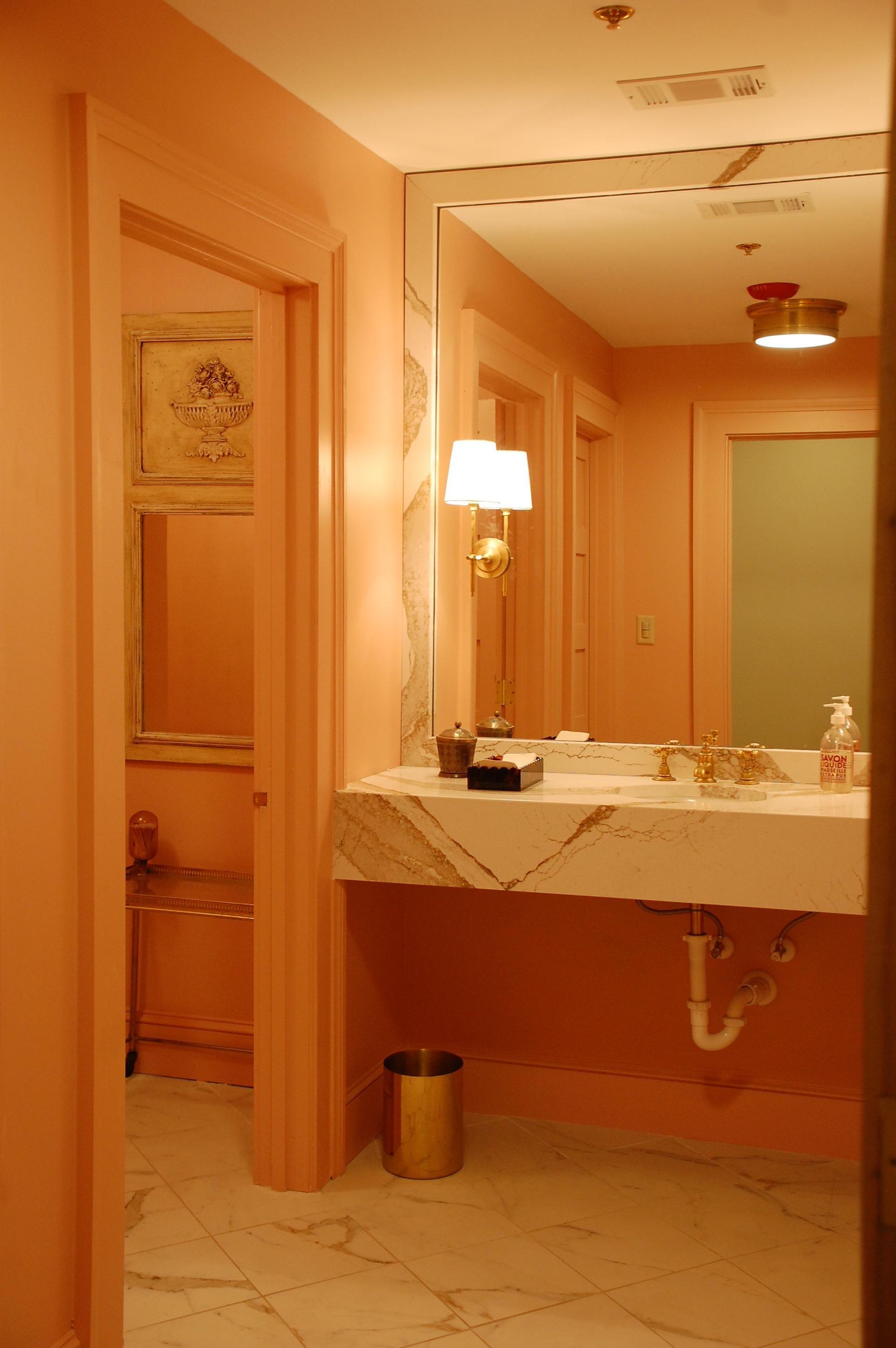 A marble-topped vanity with a sink in a peach-colored bathroom, featuring an open doorway and wall-mounted lighting.