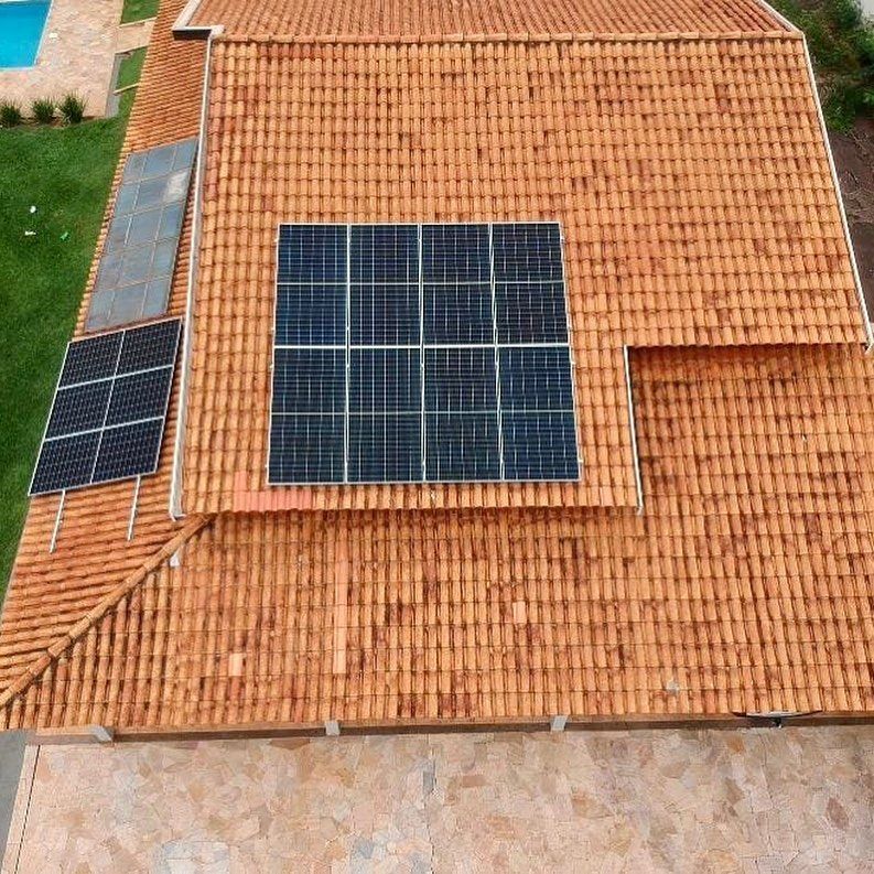 Vista de cima de um telhado de telhas laranja com dois conjuntos de painéis solares pretos instalados, ao lado de um gramado.