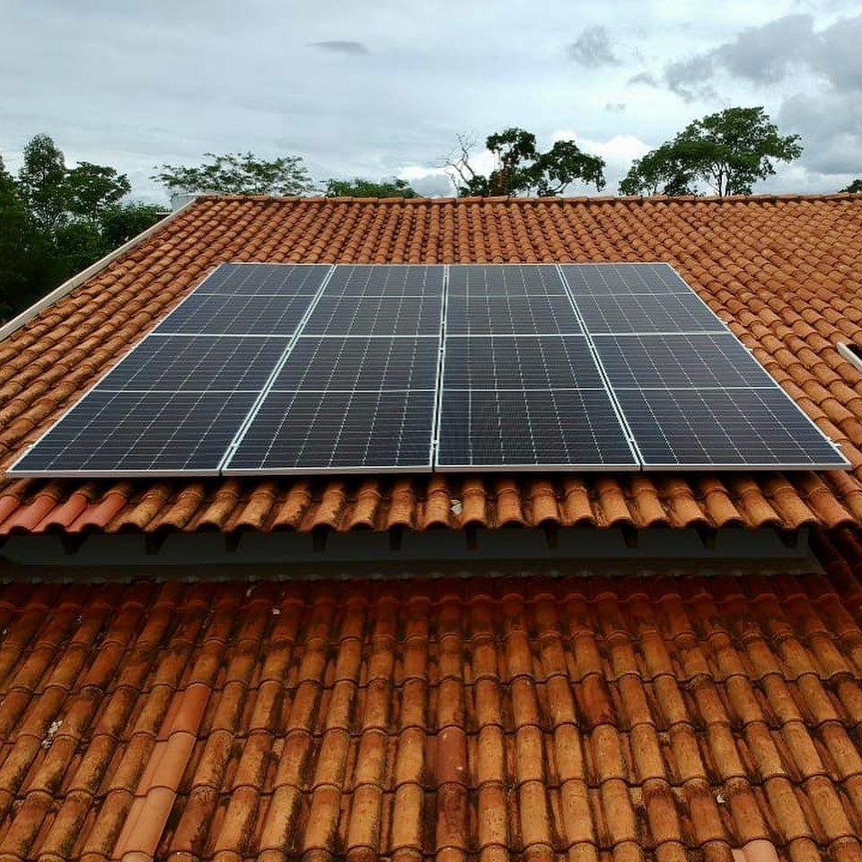 Uma matriz retangular de seis painéis solares escuros, montada no telhado de telhas de terracota de uma residência, sob um céu nublado.
