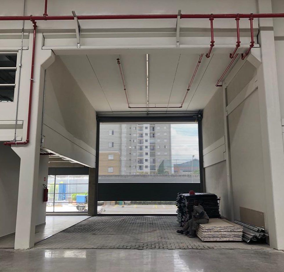 Uma doca de carga aberta em um armazém, com uma porta industrial parcialmente abaixada, revelando a vista de um prédio do outro lado da rua.