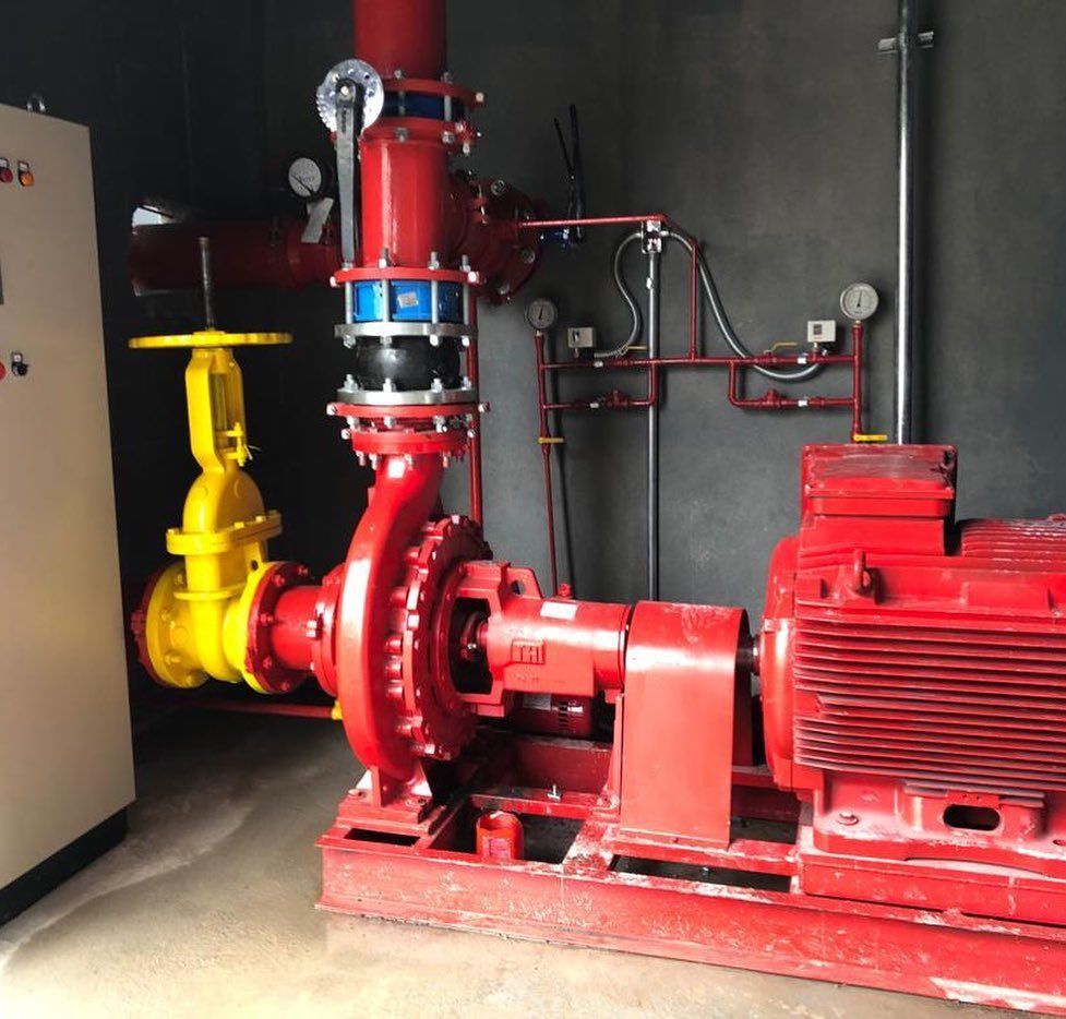 Sistema de bomba de incêndio industrial com motor e tubulação vermelhos, válvula amarela e painel de controle em uma sala interna.