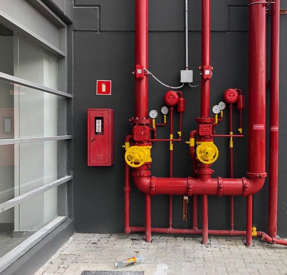 Um sistema de sprinklers contra incêndio com tubos vermelhos, válvulas amarelas e manômetros em uma parede externa cinza-escura.