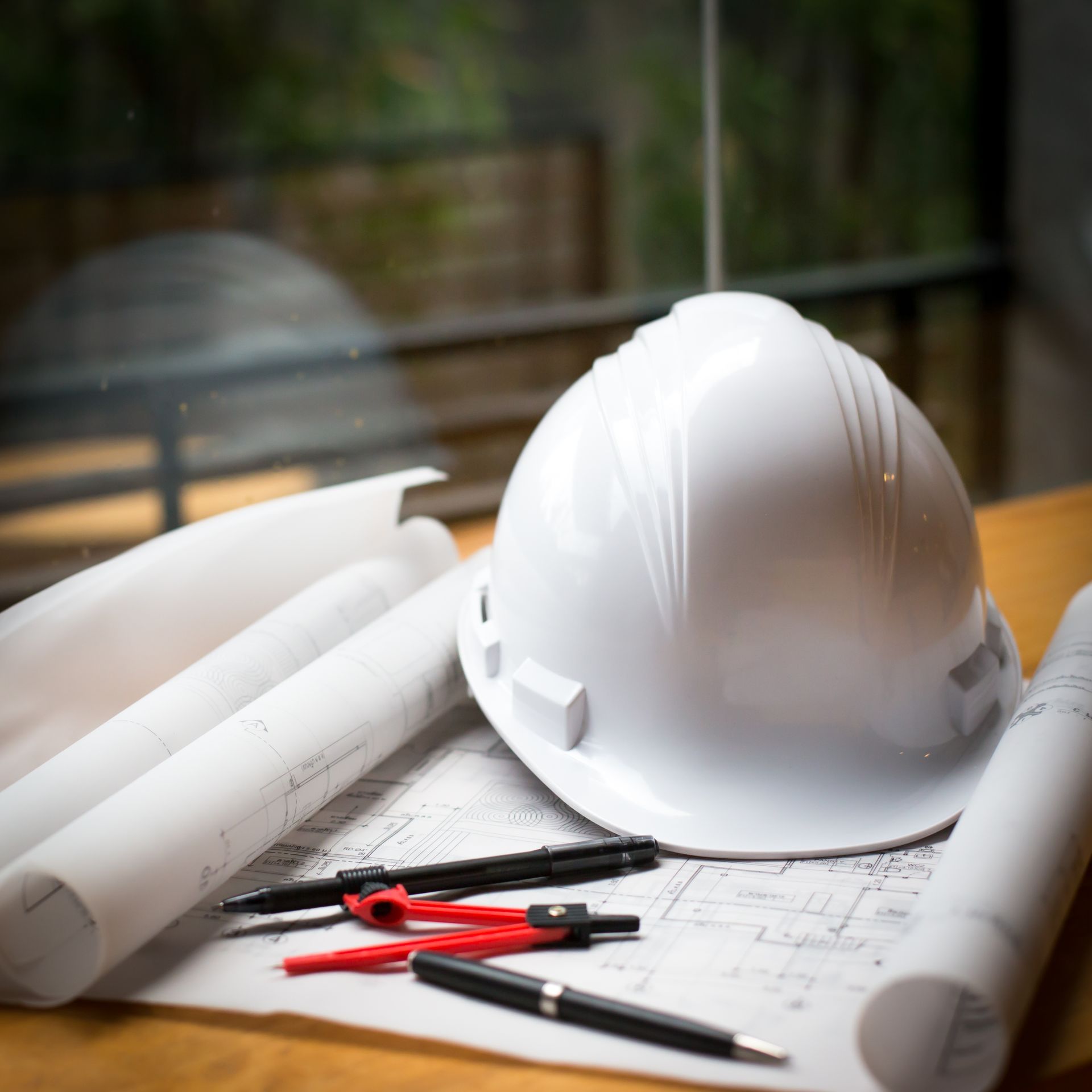 Um capacete branco, ferramentas de desenho e plantas arquitetônicas enroladas sobre uma superfície de madeira.