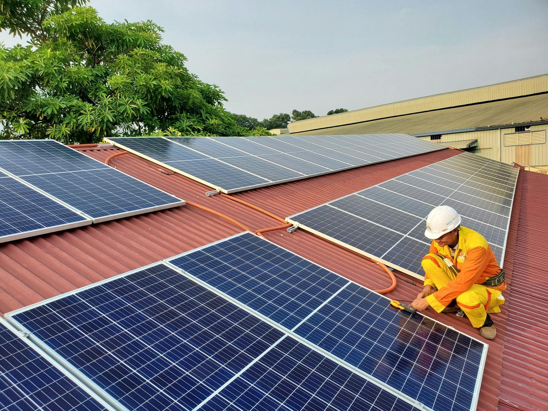 Um operário com uniforme amarelo e laranja e capacete branco realiza manutenção em painéis solares instalados em um telhado vermelho.