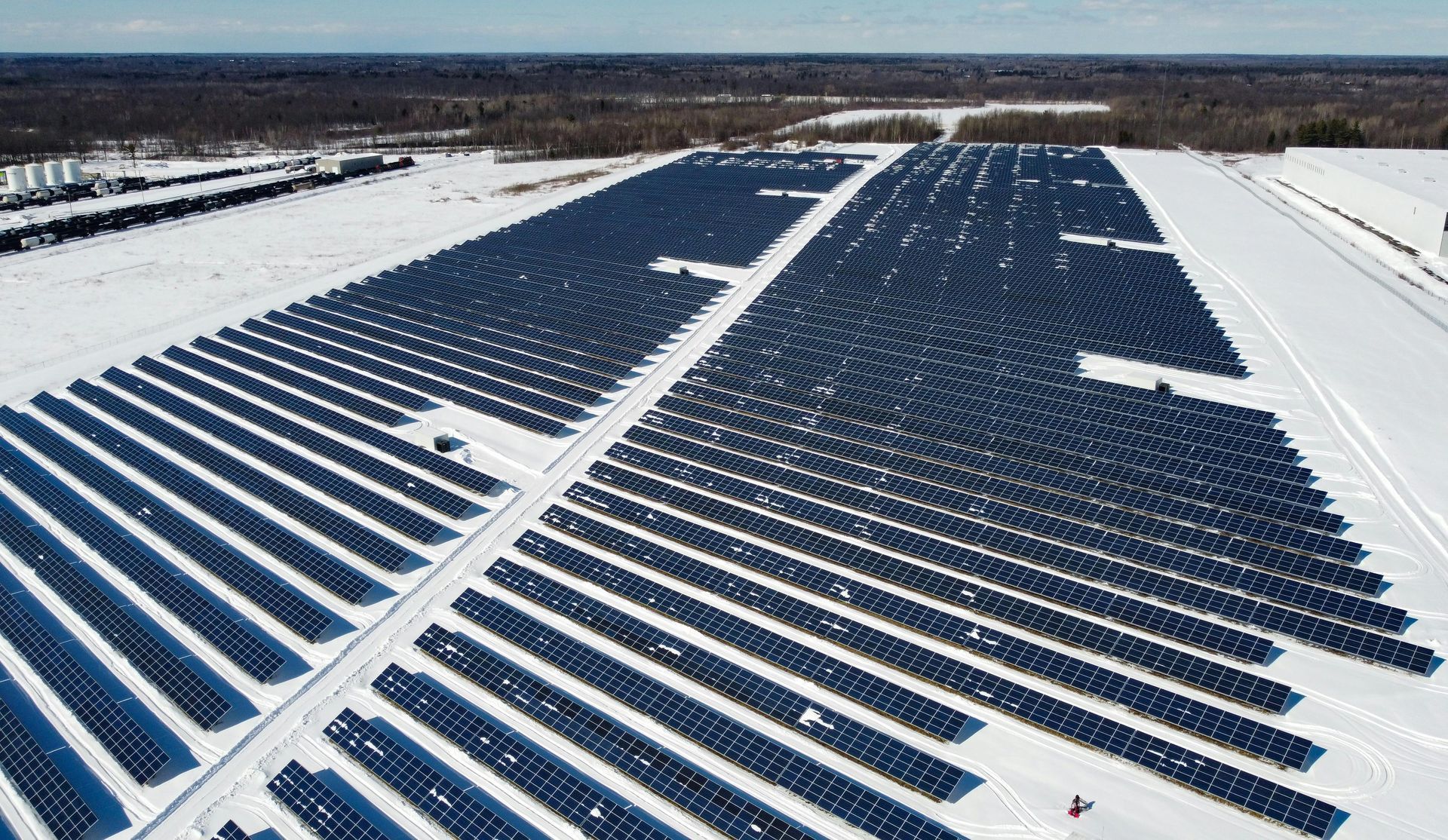 Vista aérea de longas fileiras paralelas de painéis solares escuros cobrindo um campo coberto de neve sob um céu azul claro.