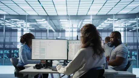 Three people working at desks in a glass-walled office, facing computer monitors.