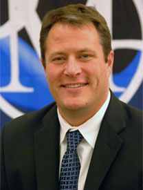 Smiling man in suit and tie standing before a blue-and-white logo backdrop