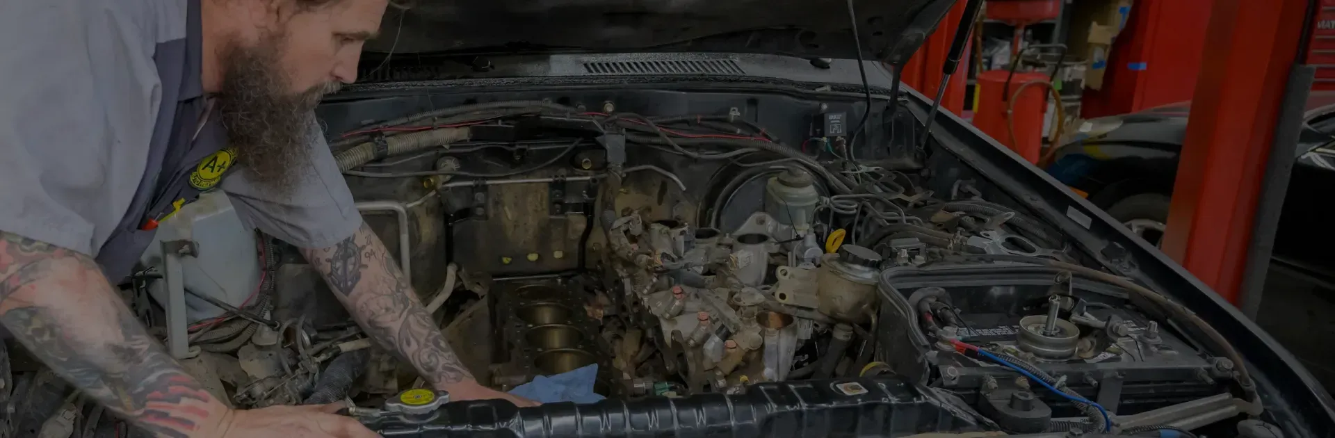 Mechanic working on a car engine in a garage, hood open, tools visible.