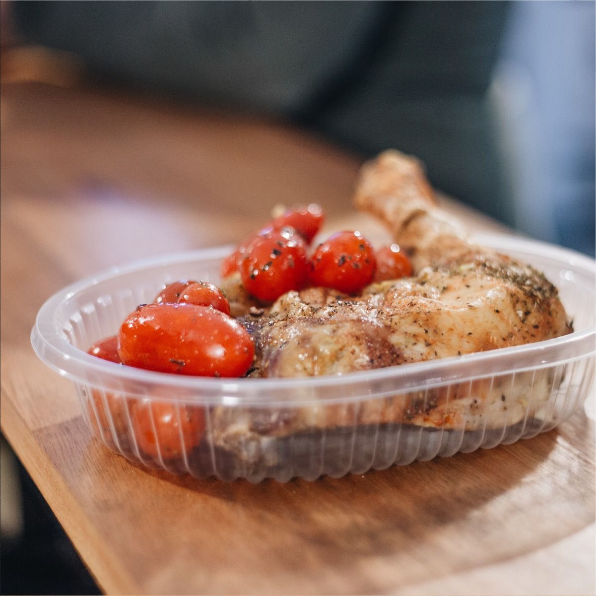 Muslo de pollo y tomates asados en un recipiente de plástico transparente sobre una mesa de madera.