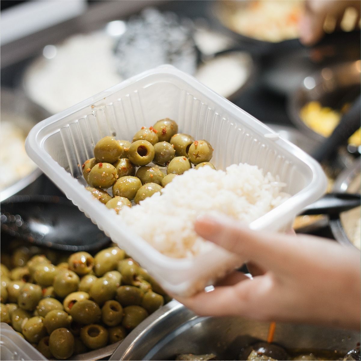 Mano sosteniendo un recipiente de plástico con aceitunas y arroz. Otros recipientes de comida al fondo.