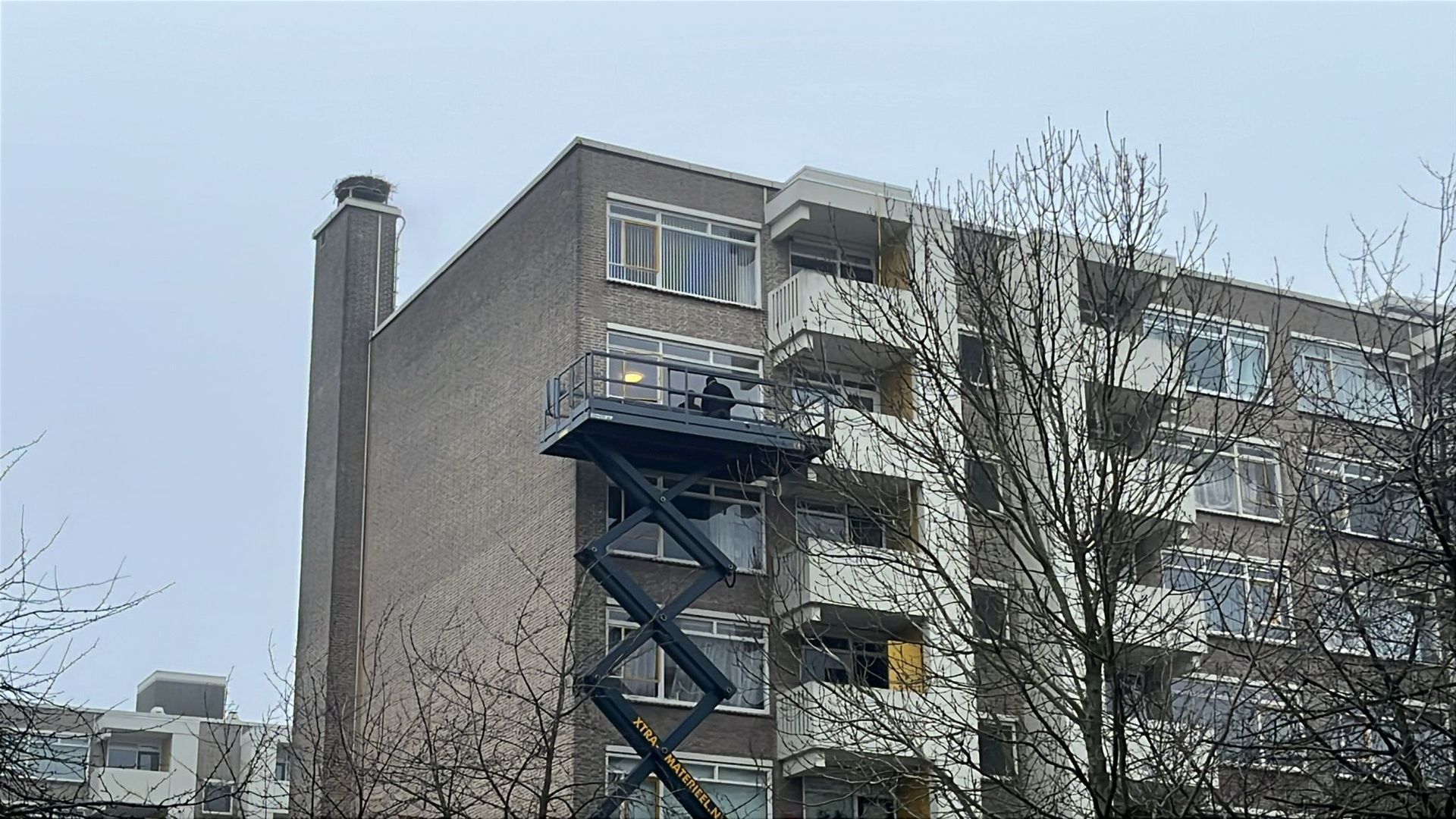 Een schaarhoogwerkerplatform schuift uit naar een bakstenen gebouw. Een persoon staat op het platform. Schoorsteen. Bewolkte dag.