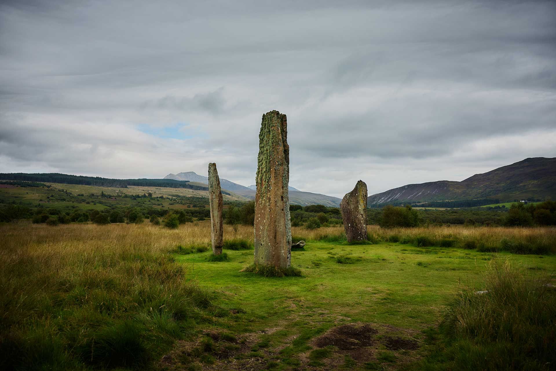 Ancient standing stones in a green field under a cloudy sky, with hills in the distance