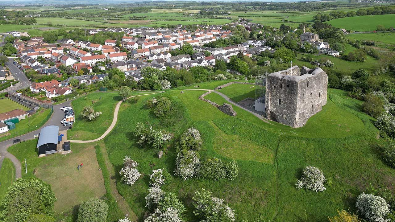 Aerial view of a village beside green fields and a large stone ruin on a hillside