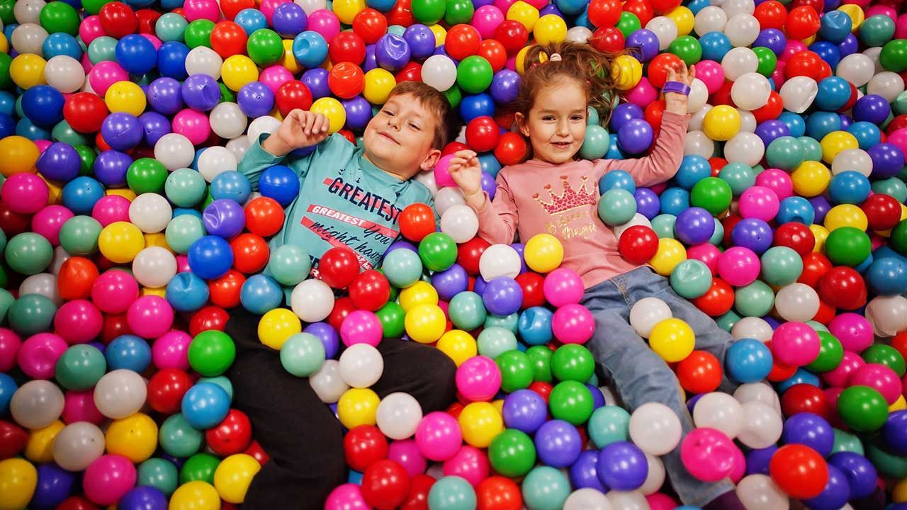 Two children lying in a colorful ball pit, smiling amid many bright plastic balls.