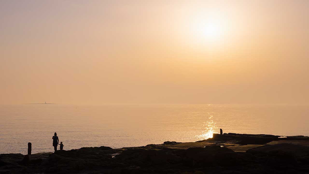 Sunset over calm sea with silhouetted people on rocky shore and bright reflection on the water