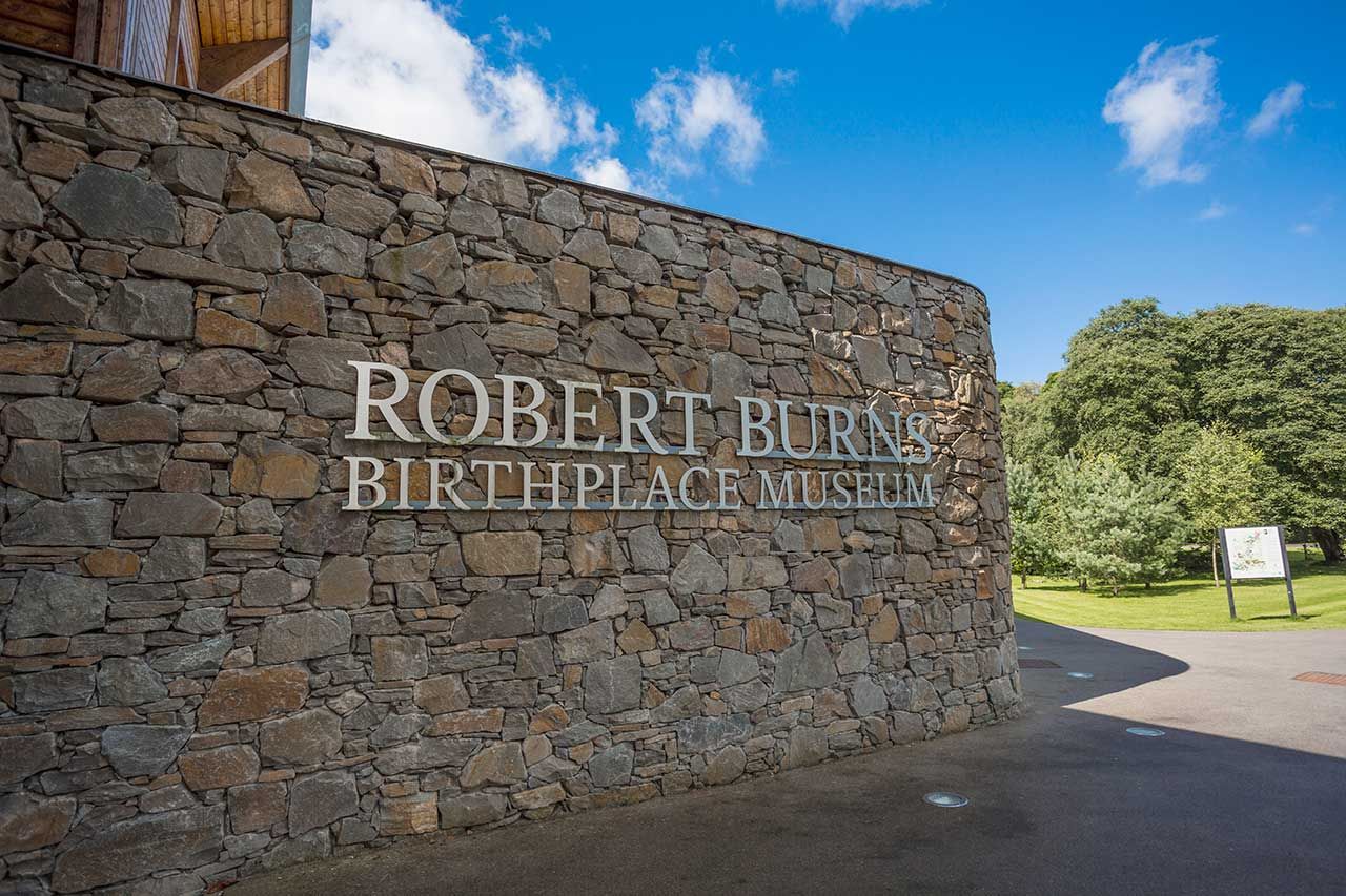 Stone wall with Robert Burns Birthplace Museum sign under a blue sky