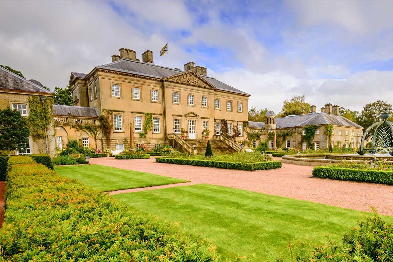 Dumfries house with manicured lawns, gravel drive, and trees under a partly cloudy sky