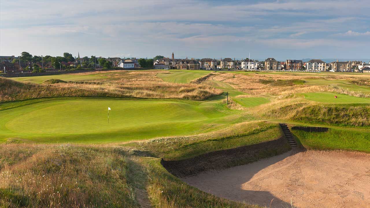Golf course green with sand bunkers and a distant row of houses under clear sky