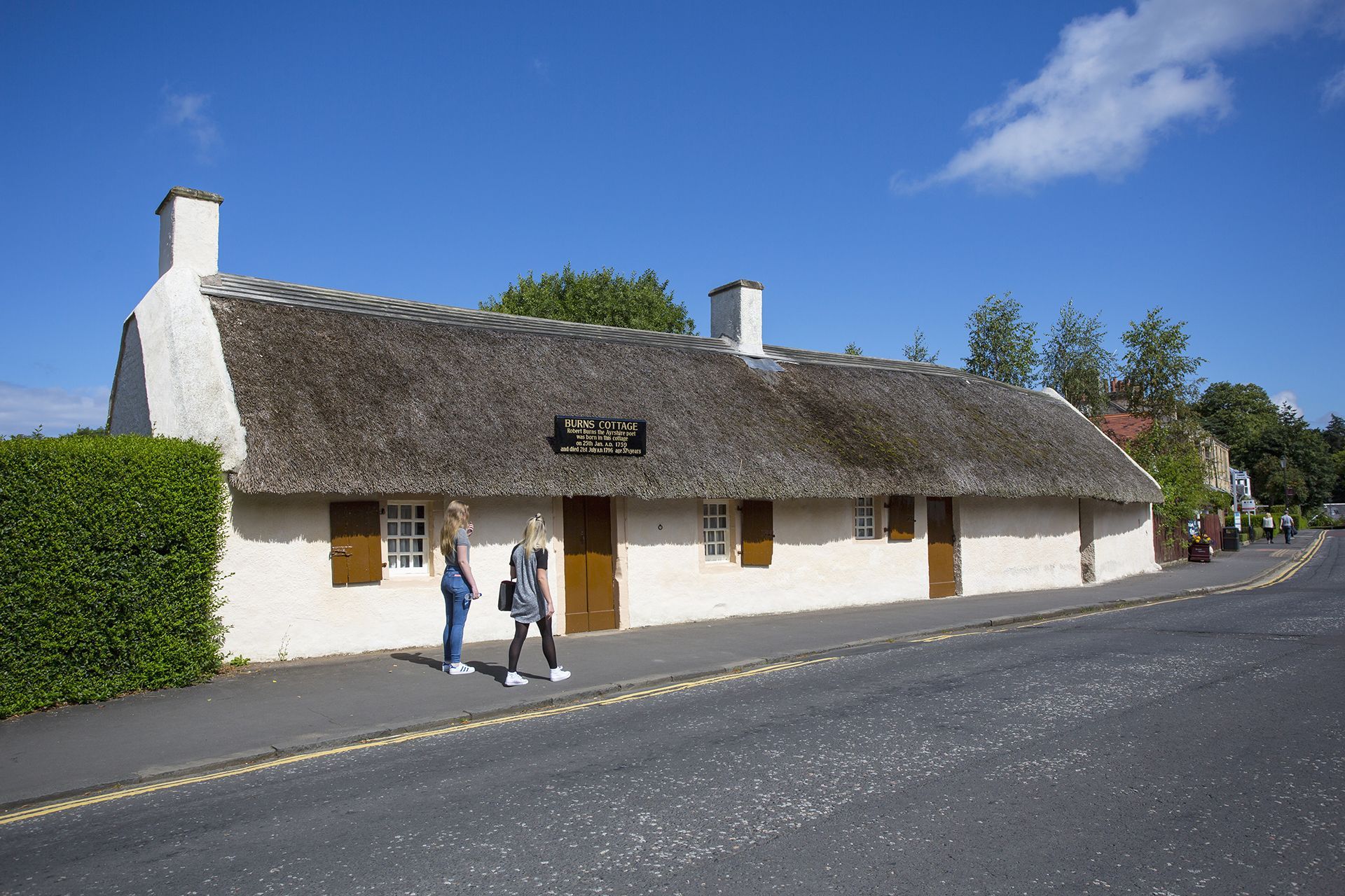 White cottage with thatched roof beside a road, with two people walking past under a blue sky