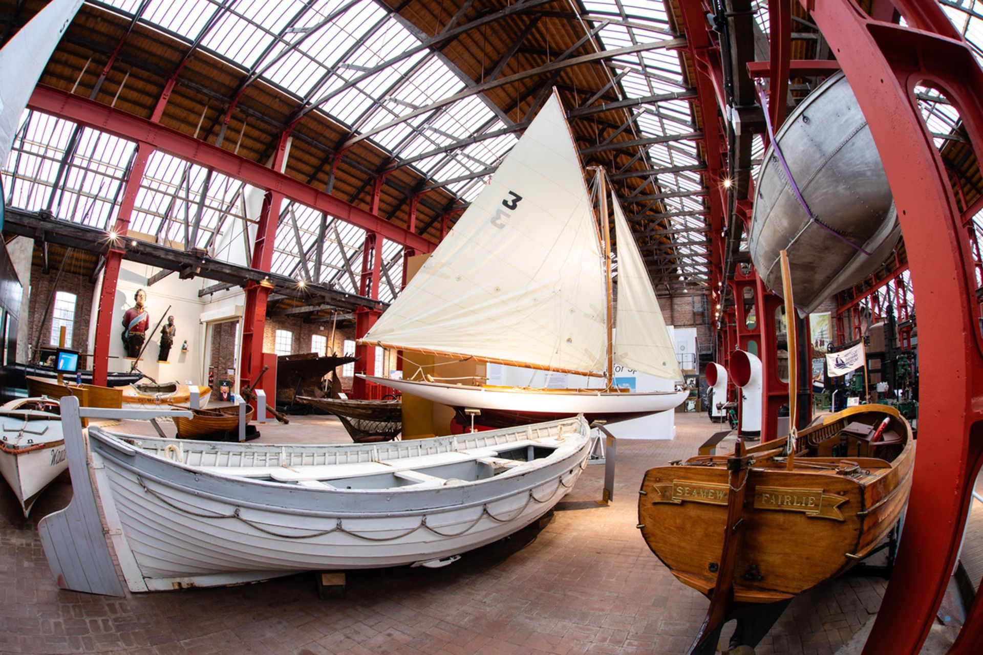 Boats with white sails displayed inside a large industrial-style museum or boathouse.