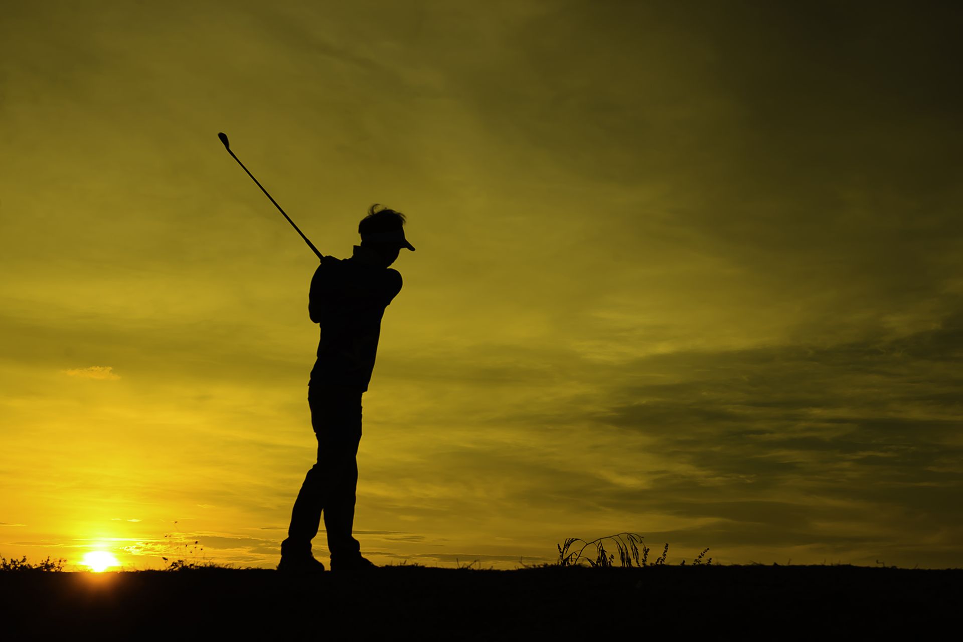 Silhouette of a golfer swinging at sunset on a hilltop, with a golden sky and low sun on the horizon.