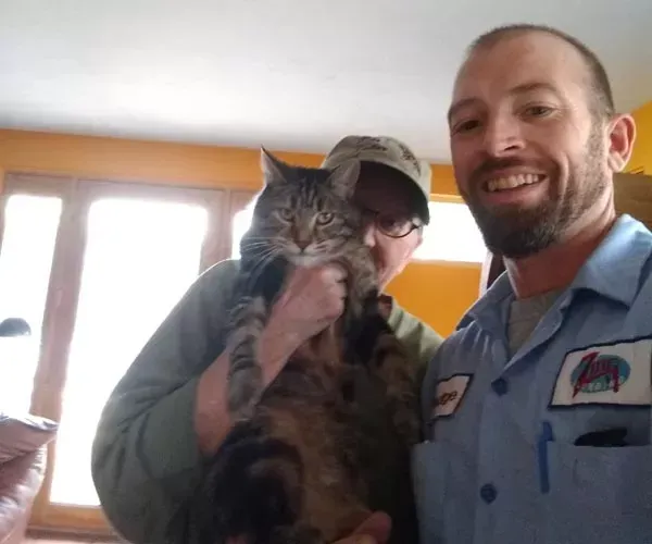 Man holds large tabby cat; another person stands behind them; indoor setting.