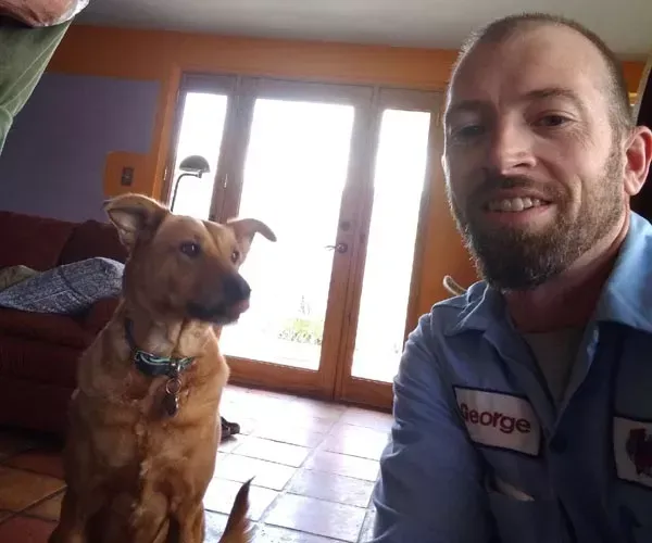 Man and brown dog smiling indoors by a door, man wearing a blue work shirt with the name 