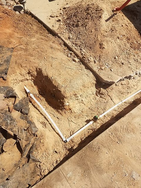 A trench in dirt with white PVC pipes laid inside. Two mounds of dirt and a concrete surface surround.