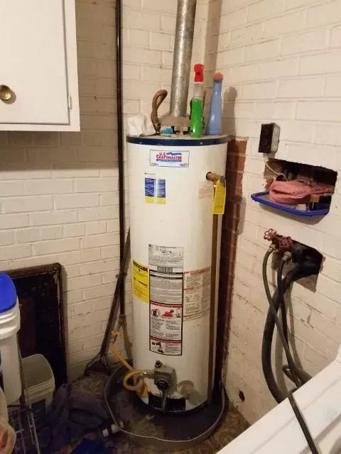 A water heater in a basement corner, next to white brick wall. It has bottles on top and exposed wires.