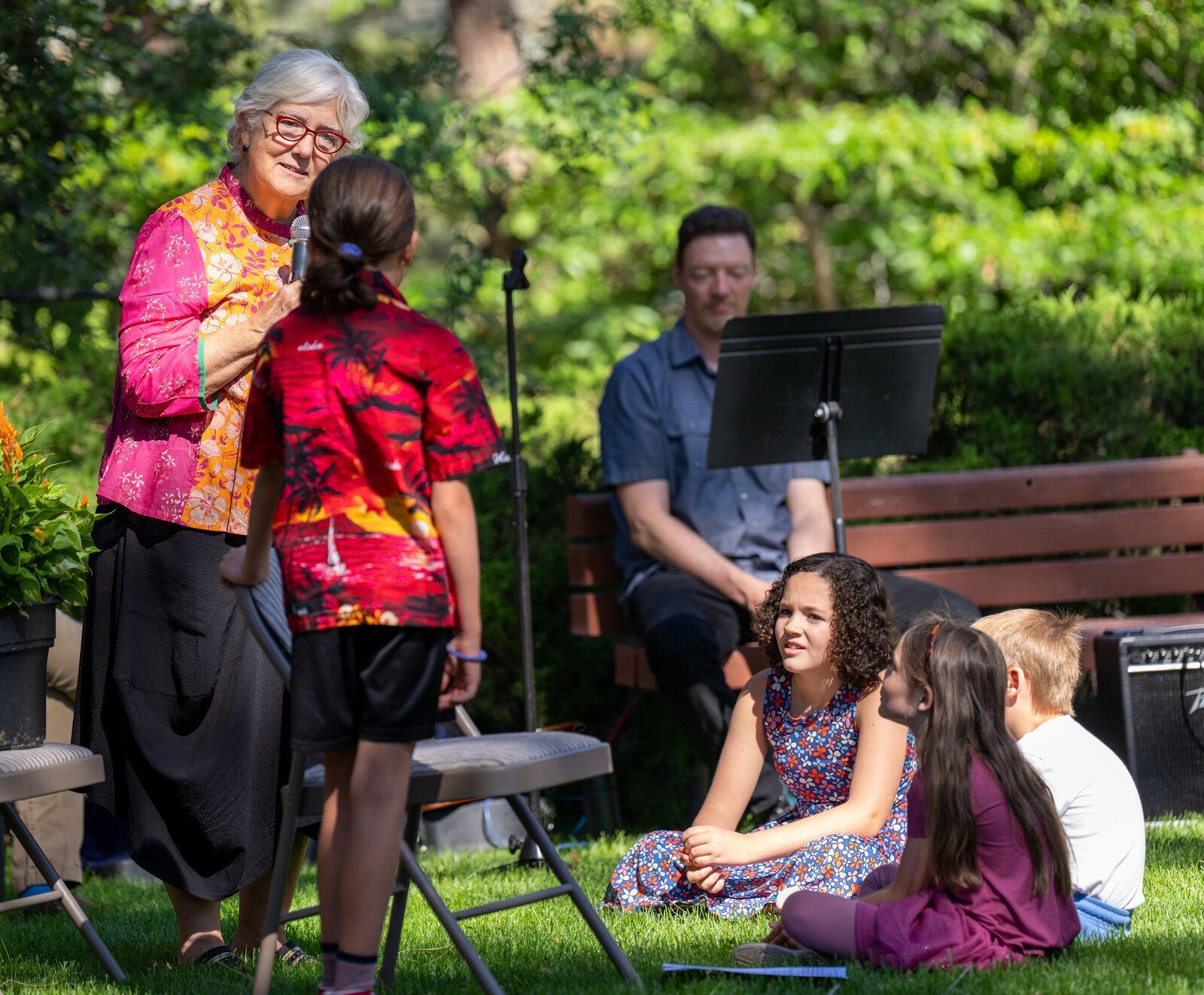 A view of the children's moment during an outdoor worship service with three children sitting on the grass looking up at a another child standing next to the pastor.