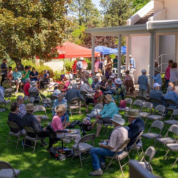 A wide view of a grassy lawn covered in chairs with people sitting in groups enjoying lunch after the outdoor worship service. Tents can be seen in the background where food is being served.