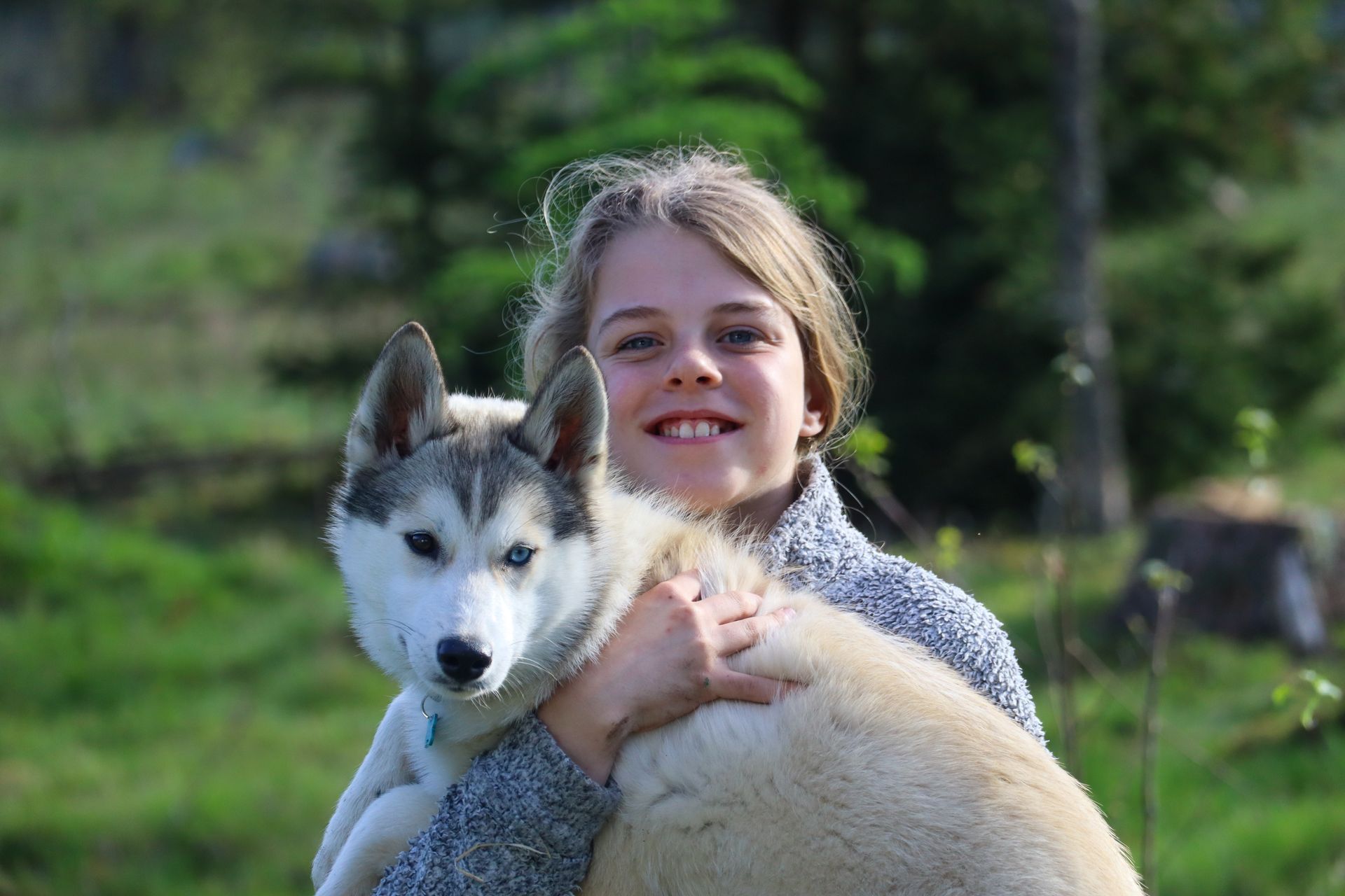 Een meisje met blond haar lacht en knuffelt buiten een husky. Groen gras en bomen op de achtergrond.