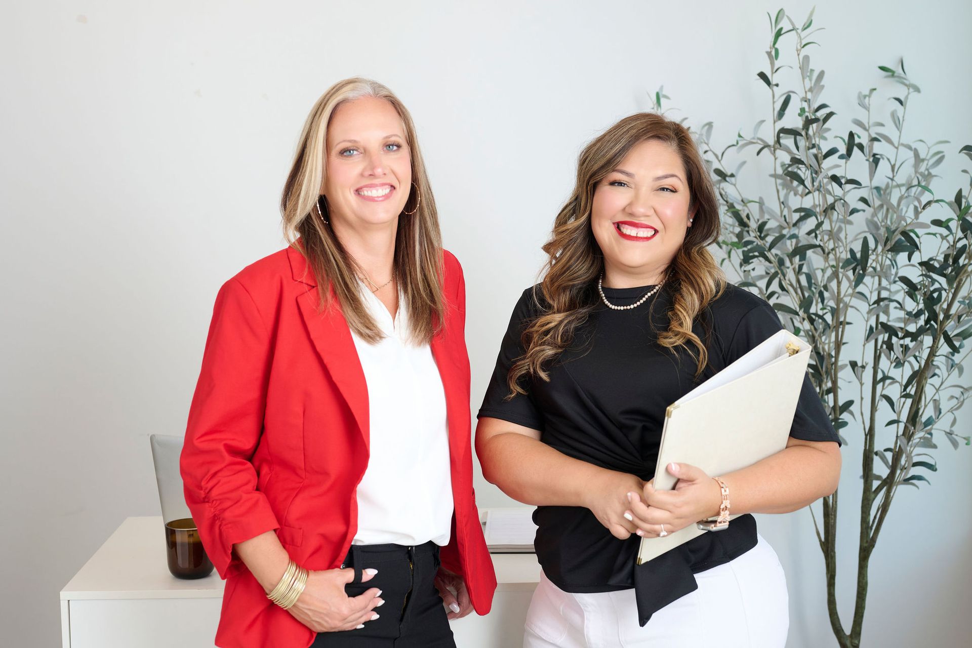 Two people smiling in an office, one holding a laptop and the other holding a folder.