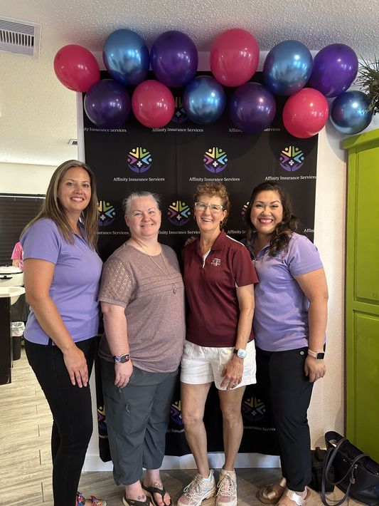 Four women posing indoors under a pink and purple balloon arch in front of a black backdrop