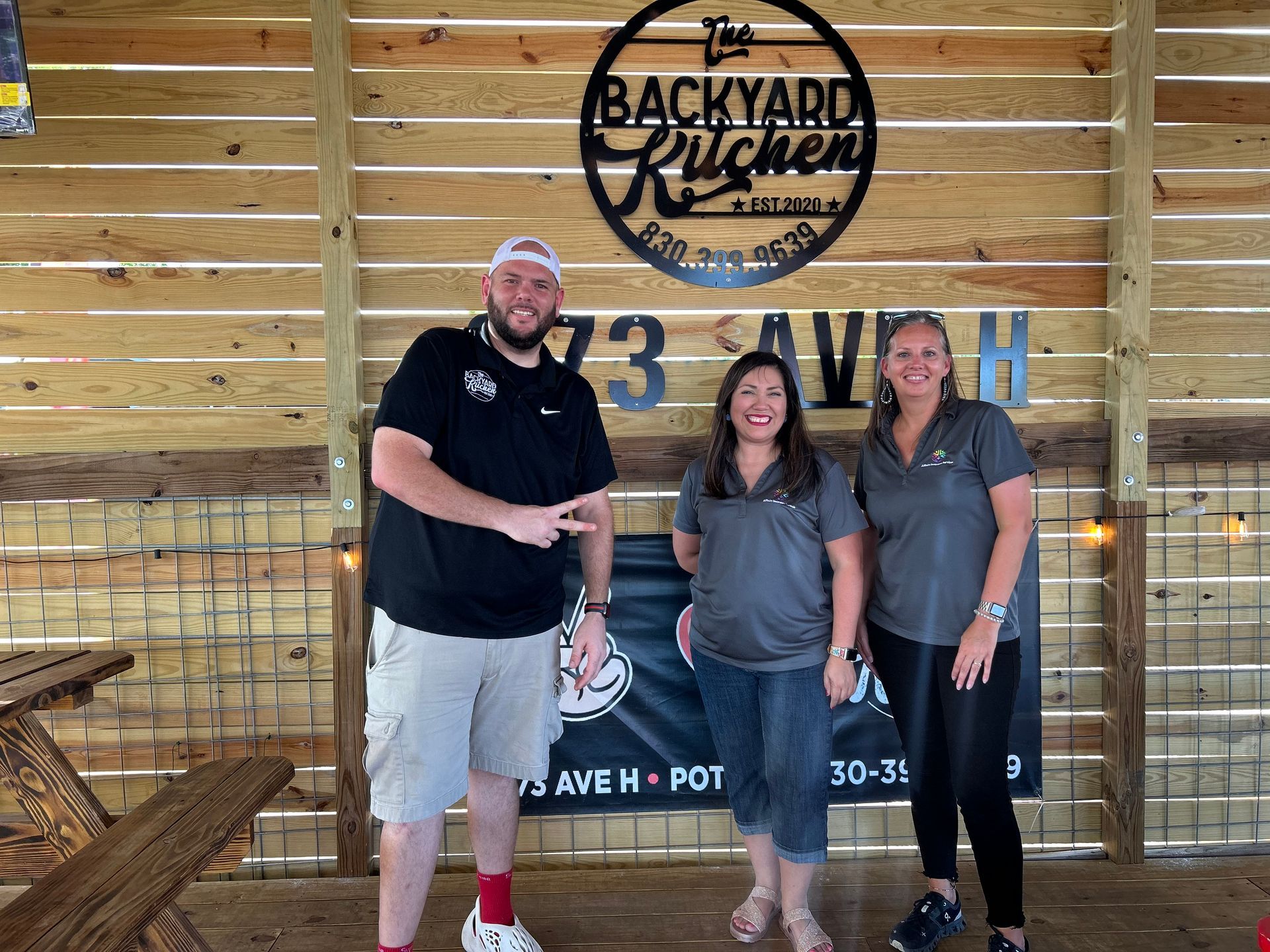 Three people posing in front of a Backyard Kitchen sign and wooden wall.