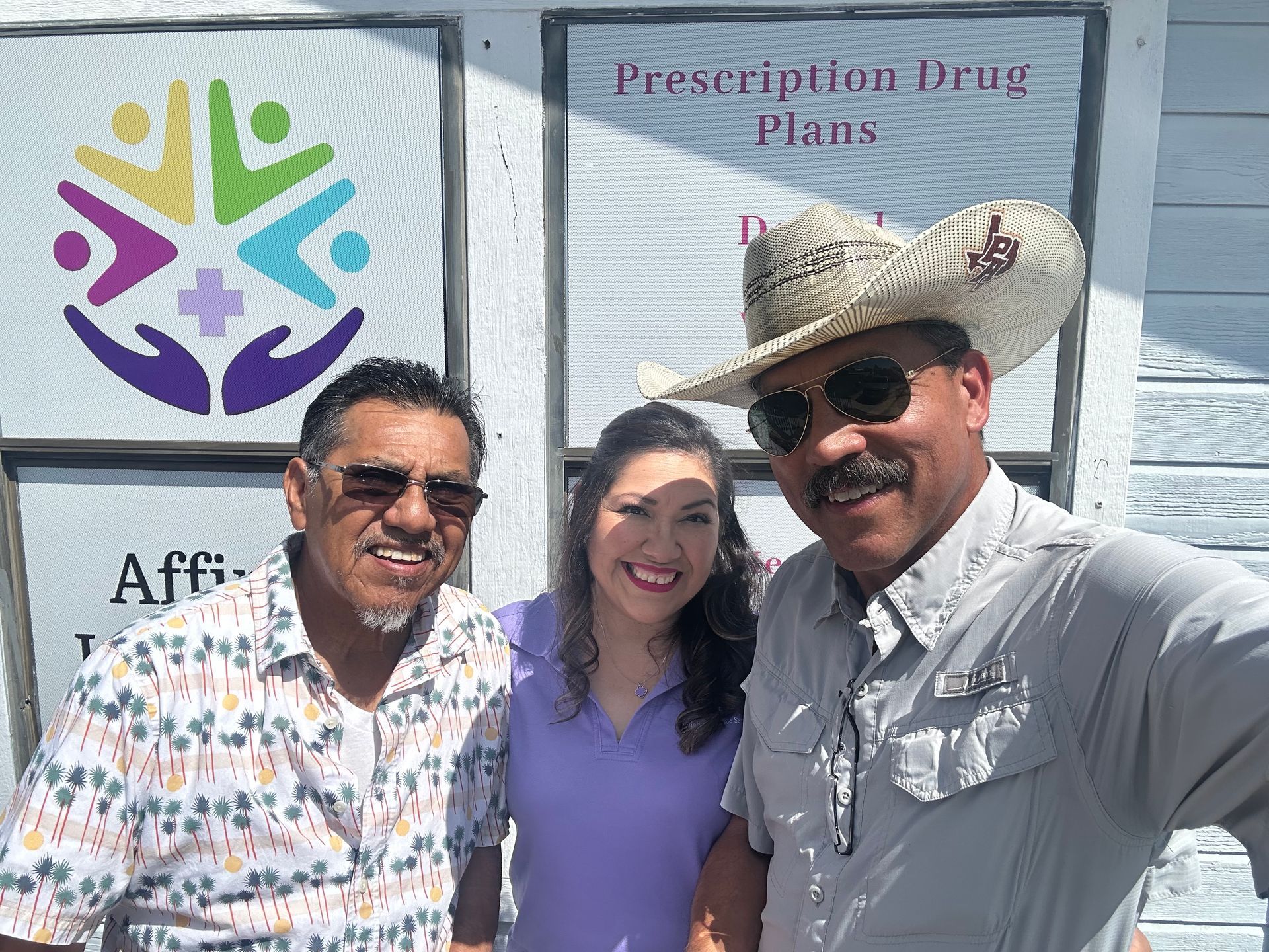 Three people posing smiling outdoors in front of a Prescription Drug Plans sign
