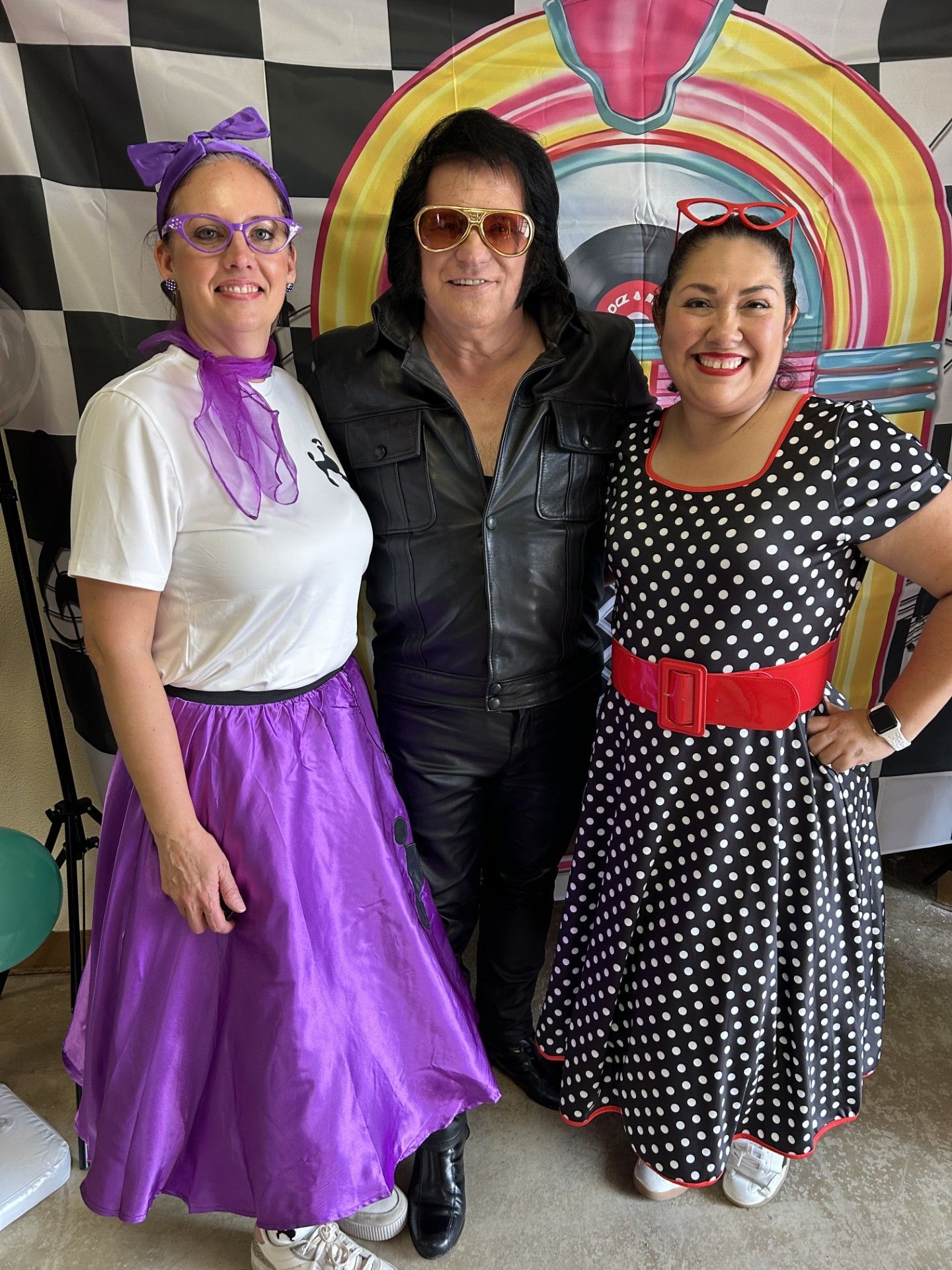 Three people pose smiling in colorful costumes, including a purple skirt and a polka-dot dress, before a rainbow mural.