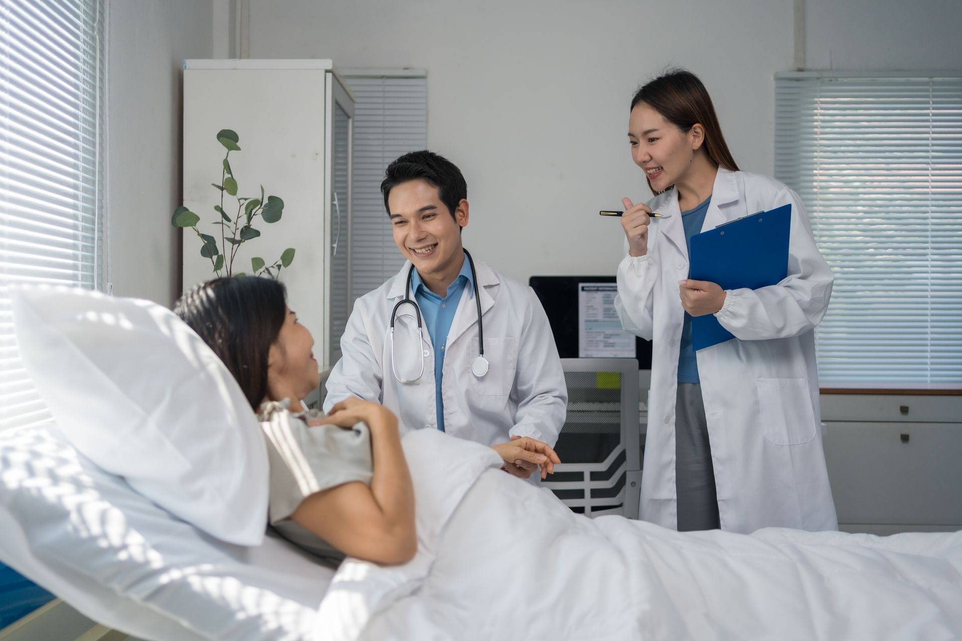 Doctor and nurse talking to a patient in a hospital bed, with the patient lying under white blankets.