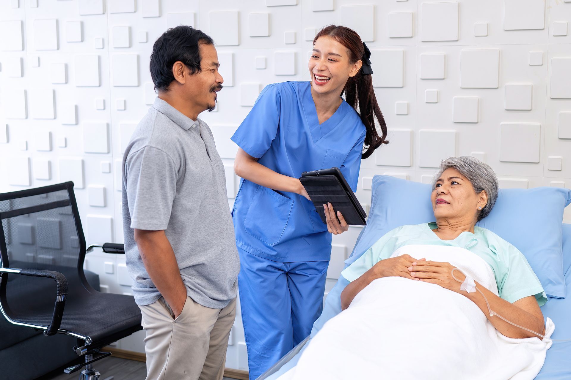 Nurse in blue scrubs talks with a man beside a smiling patient in a hospital bed