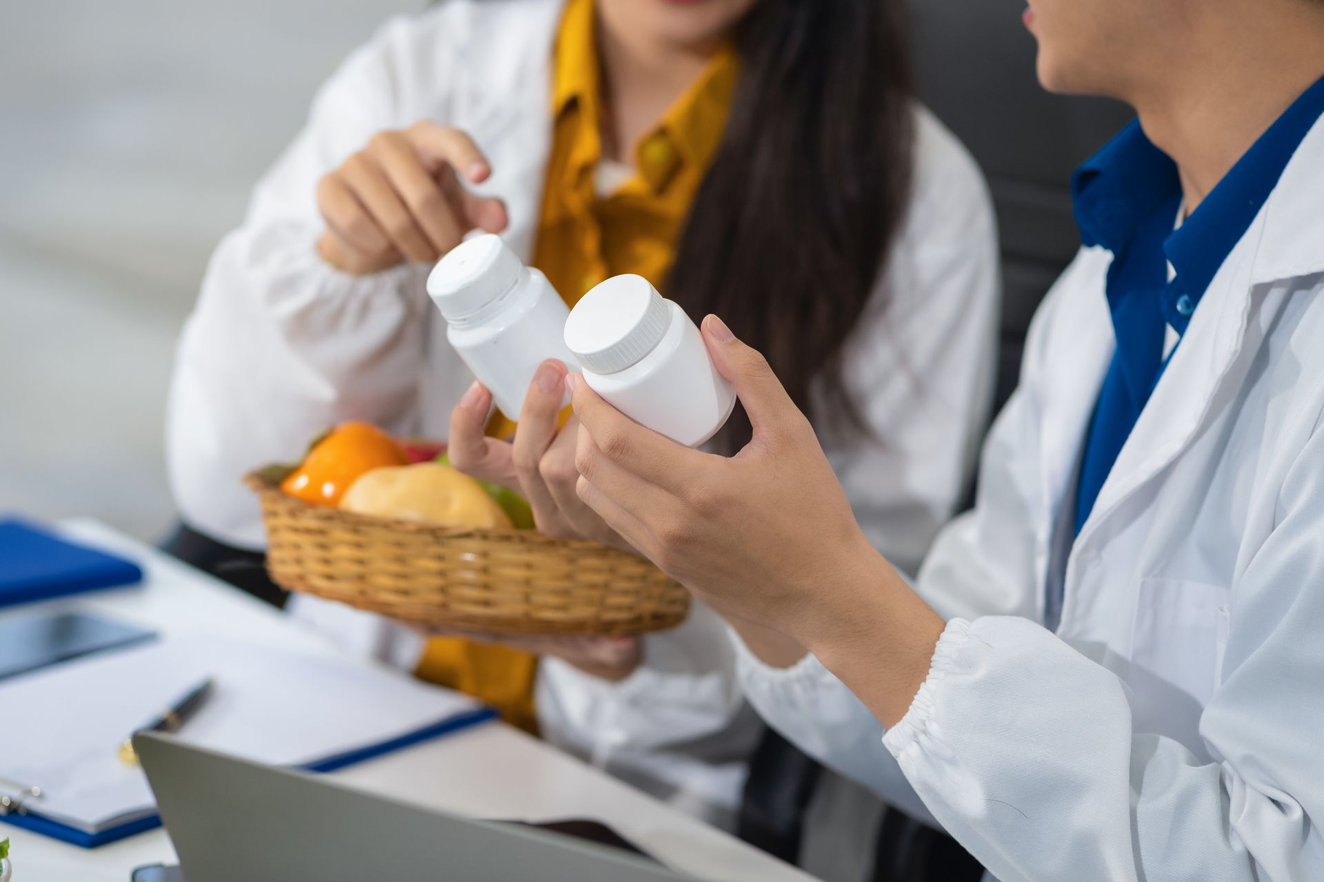Two people in white coats holding prescription bottles at a desk with a basket of oranges and a laptop.