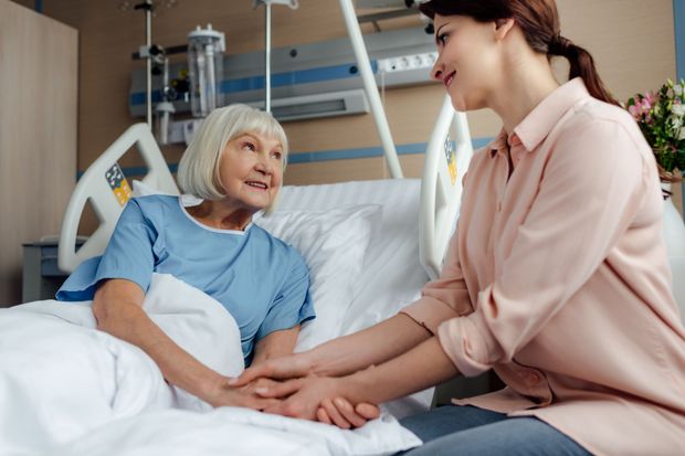 Nurse holds a patient’s hand at a hospital bedside, both smiling and talking gently.