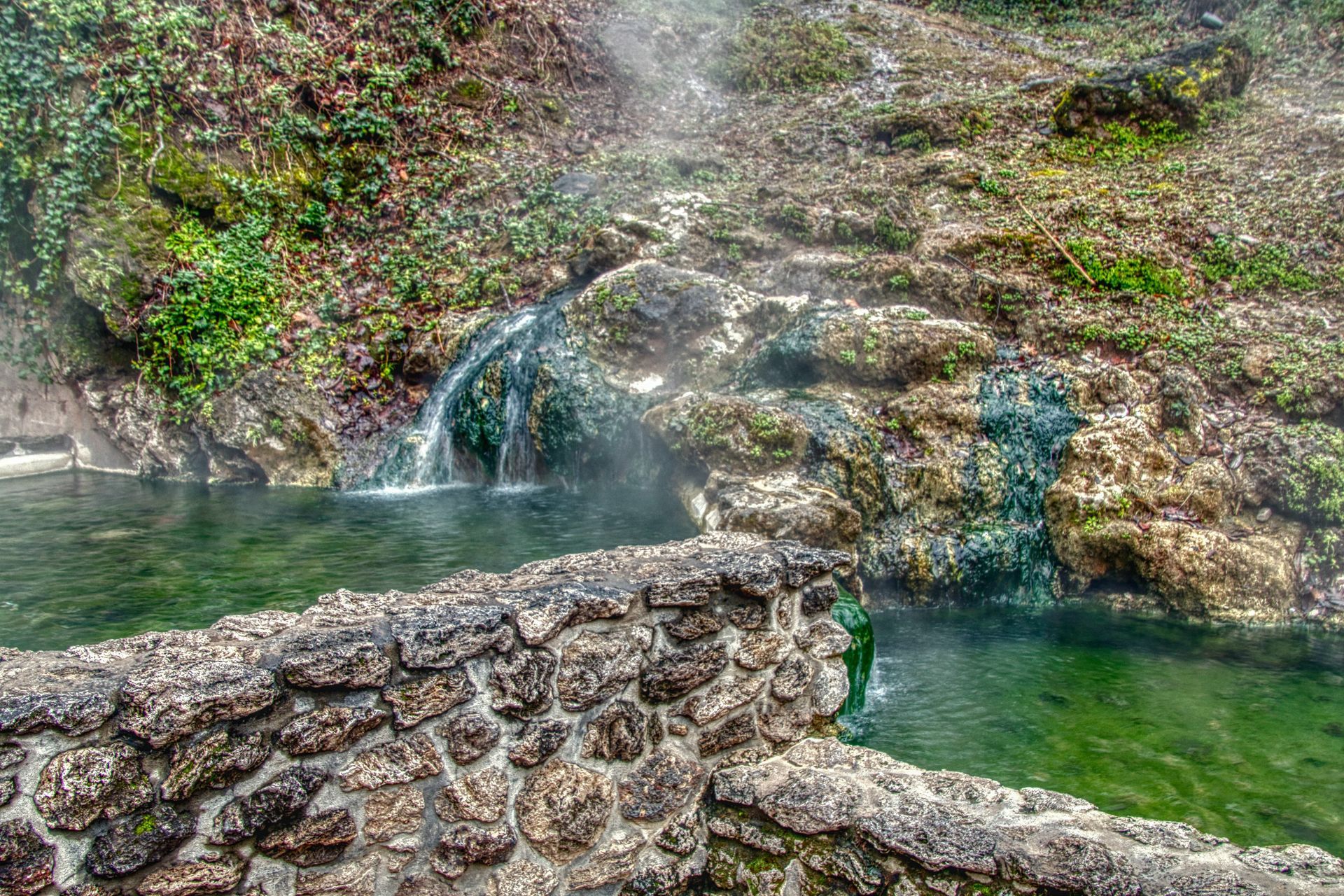 A waterfall is surrounded by rocks and trees and is surrounded by a stone wall.